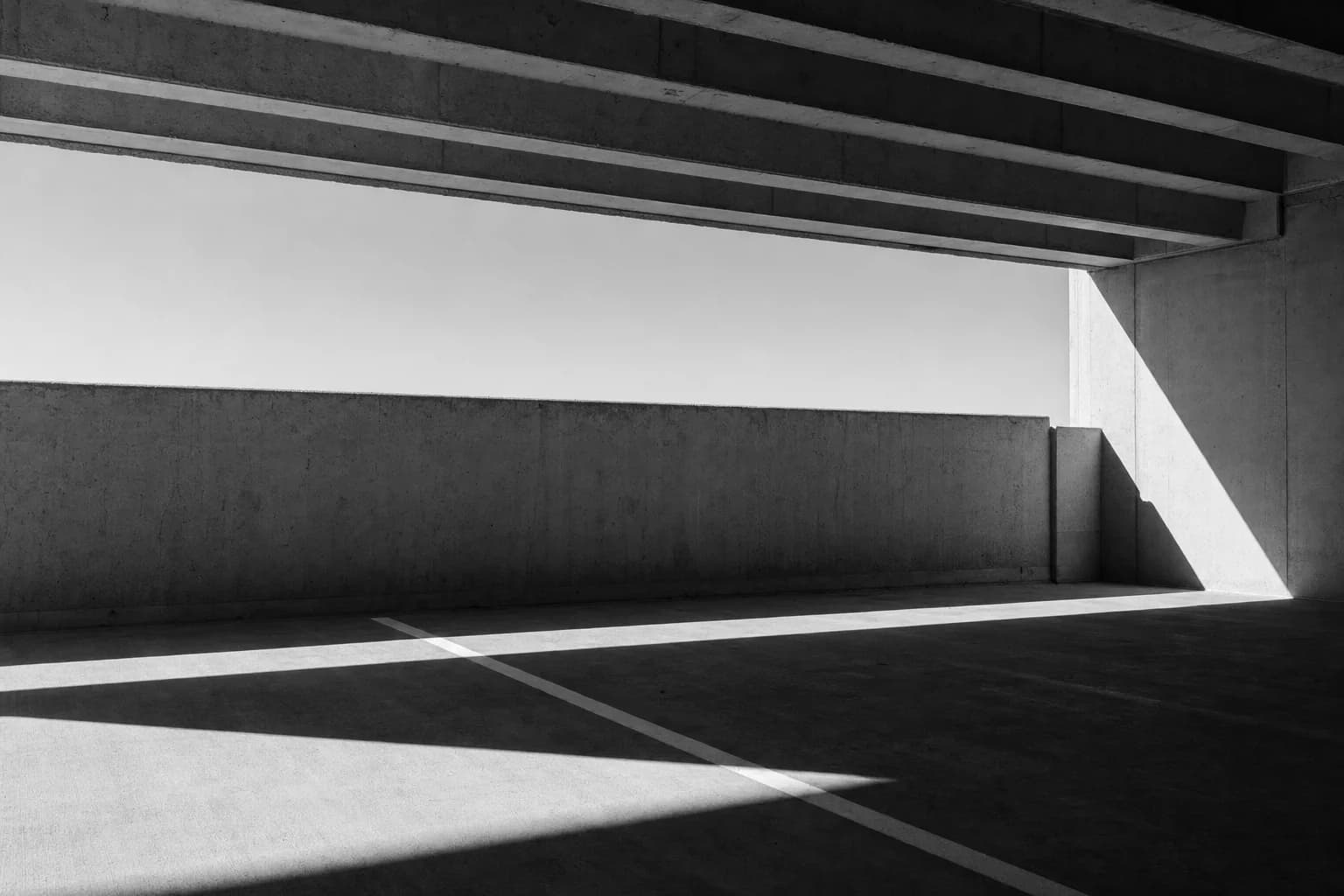 Black and white photograph of an open concrete carpark deck, beam soffit overhead and a painted bay line in raking sunlight
