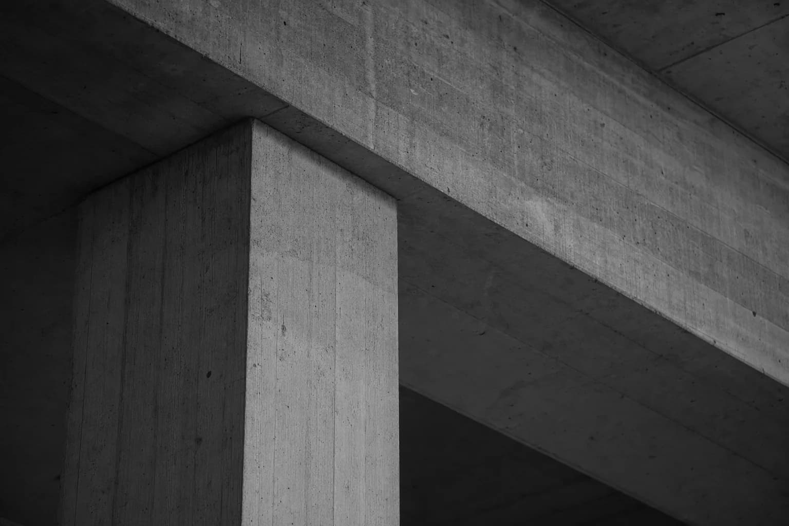 Black and white photograph of a raw in situ concrete column meeting a deep concrete beam overhead, seen from below