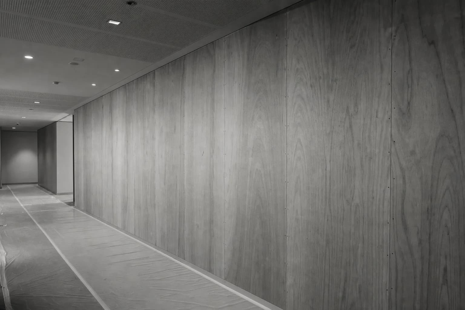 Black and white photograph of a full height plywood hoarding sealed to the ceiling with the floor finish protected up to its base